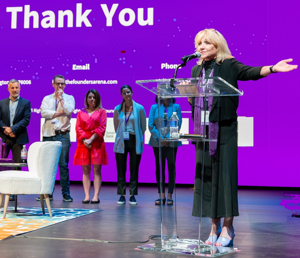 A Founders Arena leader speaking from a podium on stage with one arm extended in welcome, colleagues standing behind her.