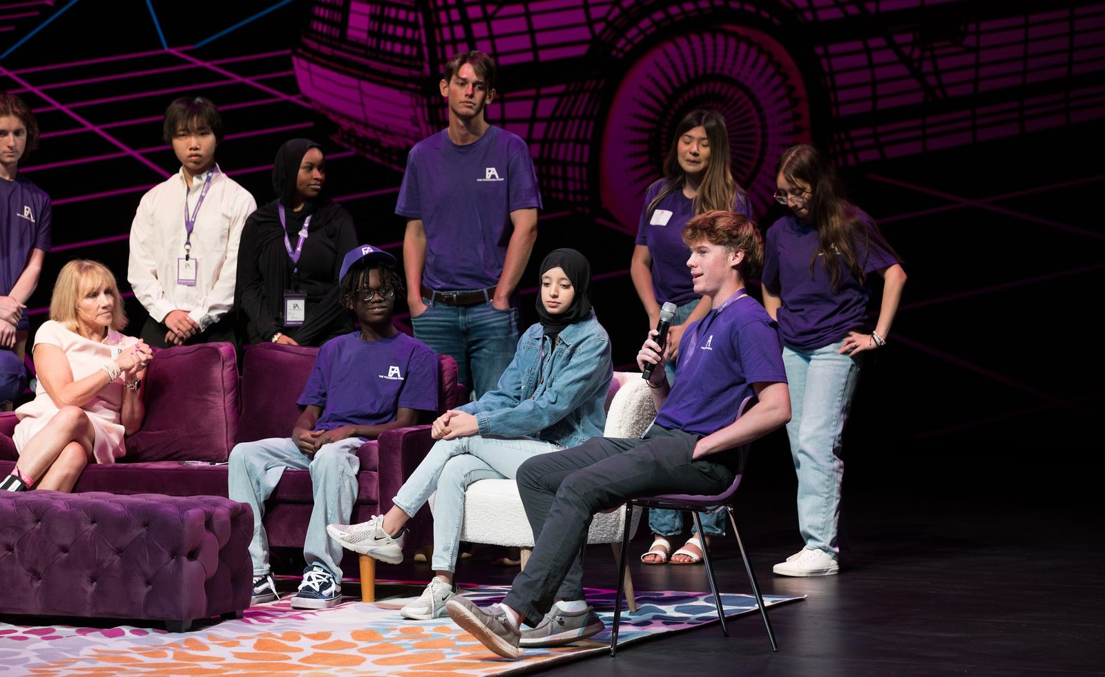 Young people in purple Founders Arena shirts on stage, one speaking into a microphone while the others listen, at a Founders Arena event.